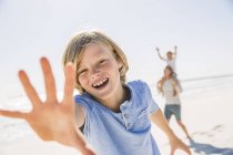 Niño en la playa mirando a la cámara, la mano levantada sonriendo - foto de stock