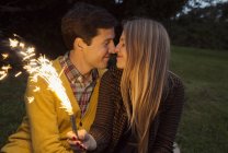 Jovem casal nariz a nariz no parque segurando fogos de artifício cintilantes — Fotografia de Stock