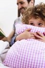 Menina abraçando travesseiro enquanto pai lendo livro de histórias na cama — Fotografia de Stock