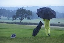 Vista posteriore del golfista accanto alla borsa da golf sotto la pioggia con ombrello — Foto stock