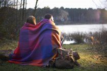 Молода пара на березі озера, загорнута в ковдру на заході сонця — стокове фото
