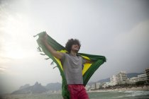 Smiling young man holding up Brazilian flag on Ipanema beach, Rio De Janeiro, Brazil — Stock Photo
