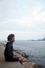 Молодой человек, сидящий на стене и смотрящий на море, Рио-де-Жанейро, Бразилия — стоковое фото