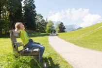 Boy sitting on park bench at rural roadside, Eckbauer bei Garmisch, Baviera, Alemanha — Fotografia de Stock