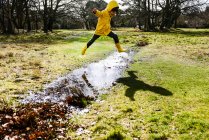 Garçon en anorak jaune sautant par-dessus flaque dans le parc — Photo de stock