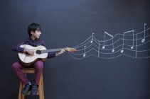Boy playing guitar in front of chalkboard wall with showing musical notation — Stock Photo