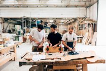 Vista frontal de los jóvenes en el taller de carpintería de pie en el banco de trabajo adjuntando ruedas al monopatín - foto de stock