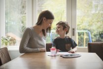 Junge lächelt Mutter an, während er am Wohnzimmertisch auf Papier malt — Stockfoto