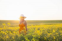 Femme adulte moyenne dans le champ de canola portant chapeau de soleil regardant loin — Photo de stock