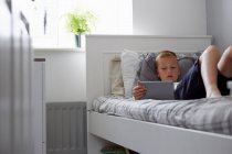 Vista a través de la puerta del niño acostado en la cama mirando hacia abajo en la tableta digital - foto de stock