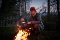 Femme mûre et fils griller des guimauves sur le feu de camp de jardin au crépuscule — Photo de stock
