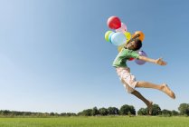Boy holding bunch of balloons leaping mid air in field — Stock Photo