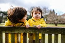 Портрет маленького мальчика и старшего брата в желтых анораках на скамейке в парке — стоковое фото