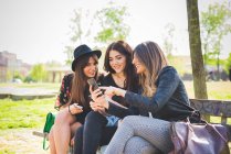 Três jovens amigas usando smartphone no banco do parque — Fotografia de Stock