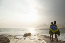 Vista trasera de dos mujeres jóvenes envueltas en bandera brasileña en la playa de Ipanema, Río de Janeiro, Brasil - foto de stock