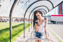Junge Frau mit Dreadlocks liest Smartphone in städtischem Wartehäuschen — Stockfoto
