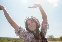 Retrato de una mujer adulta que baila en el campo usando auriculares con los brazos levantados - foto de stock