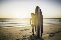 Портрет мальчика, стоящего на пляже, держащего доску для серфинга — стоковое фото