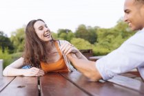 Young couple arm wrestling on picnic bench laughing — Stock Photo
