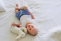 Baby girl lying on bedclothes with soft toy — Stock Photo