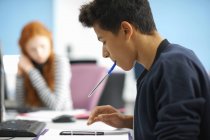 Junger männlicher College-Student am Computertisch, der auf dem Smartphone rechnet — Stockfoto