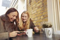 Dos amigas, sentadas al aire libre, tomando café, mirando el smartphone - foto de stock
