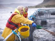 Salmon farmers feeding fish on pontoon of Scottish salmon farm over sea loch — Stock Photo