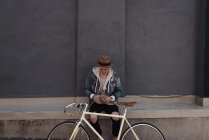 Jovem sentado na parede, de bicicleta na frente dele, usando smartphone — Fotografia de Stock