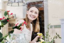 Vista através de vidro de mulher organizando flores olhando para longe sorrindo — Fotografia de Stock