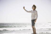 Mature man standing on beach taking self portrait using smartphone — Stock Photo