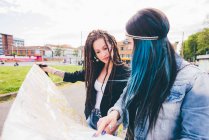Дві молоді жінки з дредлоками і пофарбованим волоссям вказують на карту в міському парку — стокове фото