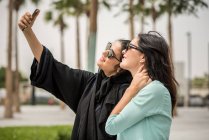 Joven mujer de mediana edad vestida con ropa tradicional usando smartphone selfie con una amiga, Dubai, Emiratos Árabes Unidos. - foto de stock