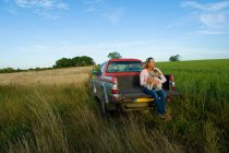 Woman sitting on back of pick up truck in a field, holding a dog — Stock Photo