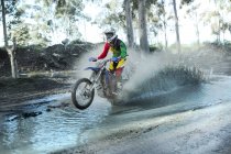 Young male motocross rider splashing through forest river — Stock Photo