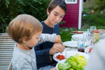 Мальчик-подросток помогает девочке есть еду на барбекю в саду — стоковое фото