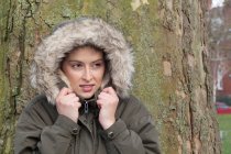 Young woman in front of park tree wrapping up in fur hood — Stock Photo