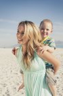 Madre dando joven hijo en piggy espalda en playa - foto de stock