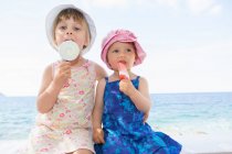 Niña y hermana usando sombreros comiendo lollies de hielo en la playa - foto de stock