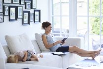 Mid adult woman using digital tablet on living room sofa whilst toddler daughter sleeps — Stock Photo
