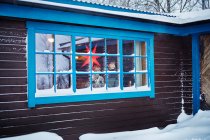 Dos hermanos mirando por la ventana de la cabaña cubierta de nieve en Navidad - foto de stock