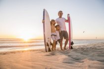 Vista completa de padre e hijos de pie en la playa sosteniendo tabla de surf al atardecer - foto de stock