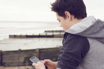 Підлітковий хлопчик на березі моря текстові повідомлення на смартфоні, Саутенд на морі, Ессекс, Великобританія — стокове фото