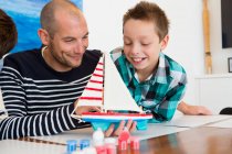 Середній дорослий чоловік і син захоплюються розфарбованим іграшковим човном за кухонним столом — стокове фото
