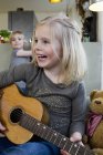 Маленькая девочка играет на акустической гитаре дома, ребенок на заднем плане — стоковое фото