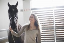Young woman leading black horse in stables — Stock Photo