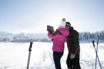 Rückansicht von Senioren-Ehepaar in verschneiter Landschaft mit digitalem Tablet, um Bergkette, Sattelbergalm, Tirol, Österreich zu fotografieren — Stockfoto