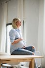 Embarazo a término mujer joven sentada en la mesa sosteniendo el estómago - foto de stock
