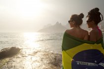 Rear view of  two young women wrapped in Brazilian flag, Ipanema beach, Rio De Janeiro, Brazil — Stock Photo