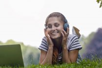 Giovane donna che indossa cuffie sdraiato su erba poggiata su gomiti guardando il computer portatile sorridente — Foto stock