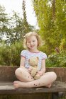 Portrait of girl on garden seat with teddy bear and star stickers on legs — Stock Photo